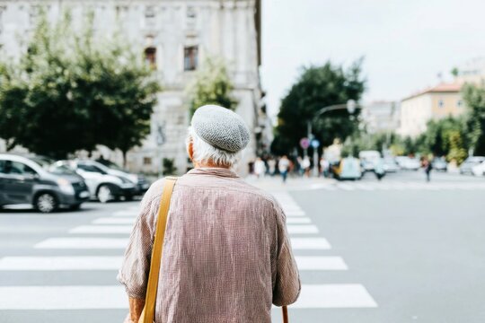 An elderly man with a cane stands at a crosswalk, carefully observing traffic in a lively city environment. Tall buildings and vehicles surround him under a clear sky - Powered by Adobe