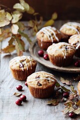Delicious muffins topped with a sweet glaze and scattered cranberries rest on a wooden table. Fall leaves add a warm touch, evoking a cozy atmosphere perfect for autumn gatherings