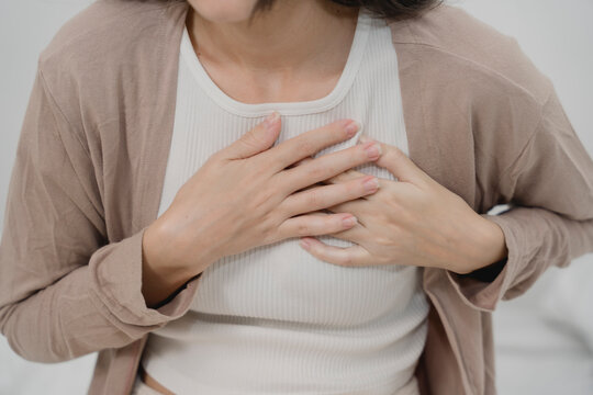 Young woman clutching her chest in pain while sitting on bed, showing signs of heart problem, chest pain, anxiety attack, or breathing difficulty, related to medical emergency symptoms.