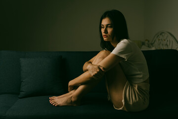 Sad young woman sitting alone on dark sofa in low light room, representing depression, loneliness,...