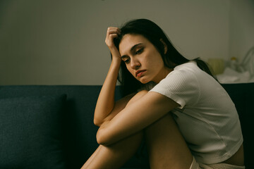 Young woman sitting alone on sofa with sad expression in dimly lit room, representing depression,...