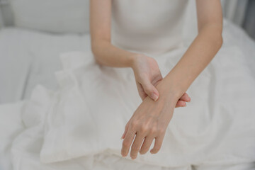 A woman sitting on bed gently massaging her wrist with a pained expression, representing wrist pain, joint discomfort, or muscle strain. Indoor setting with soft natural lighting.