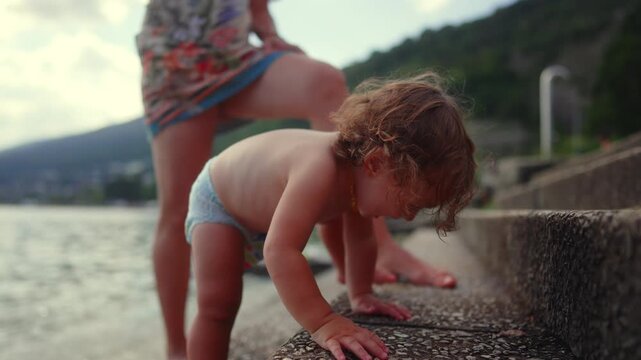Toddler crying while mother holds him back from touching stone steps near lake, frustrated wanting to explore textured surface, emotional moment of curiosity, control, and safety