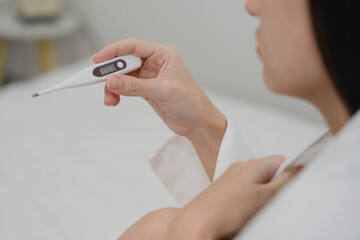 Young sick woman wrapped in blanket sitting on bed and checking thermometer, showing symptoms of...