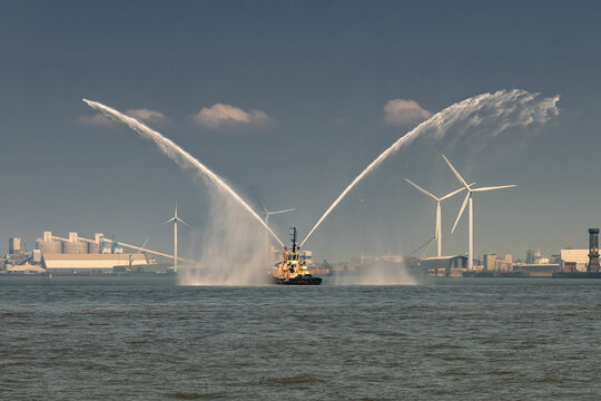 A tugboat in Liverpool sprays water into the air as part of a maritime celebration, with wind turbines and industrial buildings in the background.