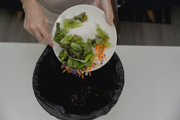 Woman scraping fresh salad into trash bin, representing food waste, poor appetite, sustainability...