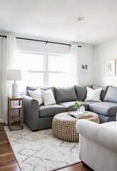 Fototapeta premium A bright and inviting living room with a comfortable gray sectional sofa, a cozy woven coffee table, and large windows creating a light-filled, airy space. 