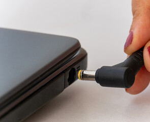 Woman's hand inserts the plug of a power supply adapter cable into the power jack of a laptop....