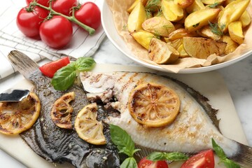 Tasty roasted flounder fish served with potatoes, tomatoes, basil and lemon slices on white marble table, closeup. Homemade seafood dish