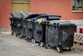 Row Of Large Black Garbage Bins With Open Lids And Waste Overflowing On Urban Street: Plastic Trash...
