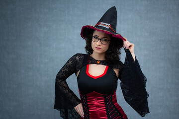 Charming witch, with curly hair, wearing eyeglasses, touching her witch's hat with one hand. Ideal for Halloween, trick or treat, fantasy, october holiday. Horizontal studio portrait with copy space.