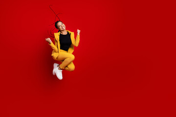 Young elegant woman jumping joyfully against a vibrant red background wearing a trendy yellow suit...
