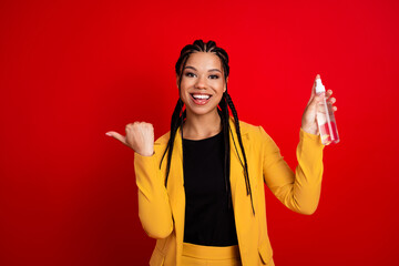 Confident young woman in a stylish yellow suit holding spray bottle and gesturing, against vibrant...