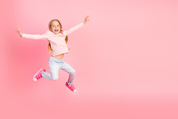 Cheerful young girl jumping with happiness in casual outfit on pink background showing carefree and...