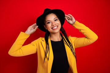Confident young woman in stylish yellow blazer with black hat, smiling against bright red...