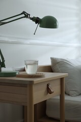 Bedside table with lamp and glass of water near bed indoors, closeup