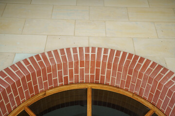 Brickwork forms a striking arch above a wooden window, showcasing craftsmanship and architectural detail. The backdrop features a smooth, light-colored stone wall, highlighting the design.