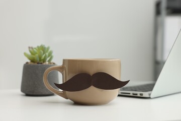 Happy Father's Day. Cup with paper moustache and laptop on white table indoors, closeup