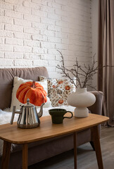 A living room that showcases a cozy sofa along with beautiful natural autumn decor elements and accents: teapot, glass, ceramic vase with dry flowers and branches, pillows with autumn pumpkins decor.
