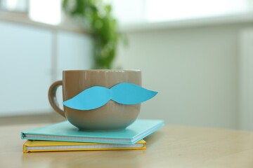 Happy Father's Day. Cup with mustache and notebooks on wooden table indoors, closeup. Space for text
