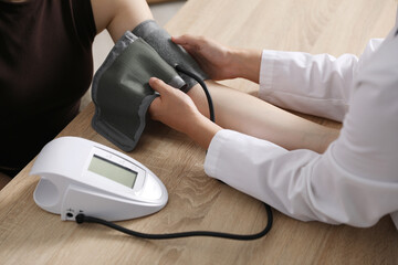Doctor checking woman's heart rate and blood pressure via sphygmomanometer at wooden table indoors,...