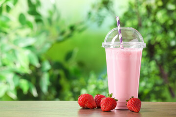 Tasty milkshake in plastic cup and strawberries on wooden table outdoors, space for text