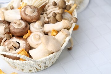Different raw mushrooms in wicker basket on white tiled table, closeup