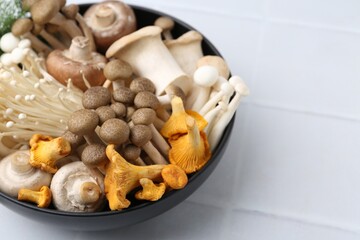 Different raw mushrooms in bowl on white tiled table, closeup. Space for text