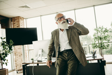 Elderly businessman in a stylish jacket engaging in a phone call while leaning on his office desk...