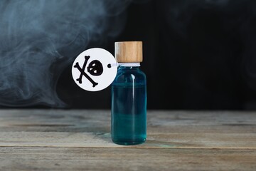 Bottle of poison with warning sign on wooden table against dark background with smoke