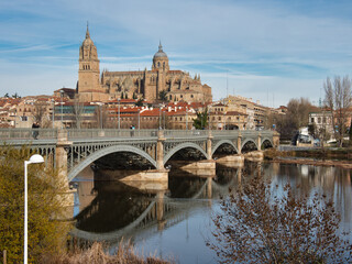 Obraz premium Salamanca Cathedral and Enrique Estevan Bridge: The iconic Salamanca Cathedral and the Enrique Estevan bridge with their reflections in the calm waters of the Tormes River on a sunny day.