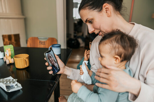 Mother showing sick son to female pediatrician while taking assistance on video call through smart phone at home