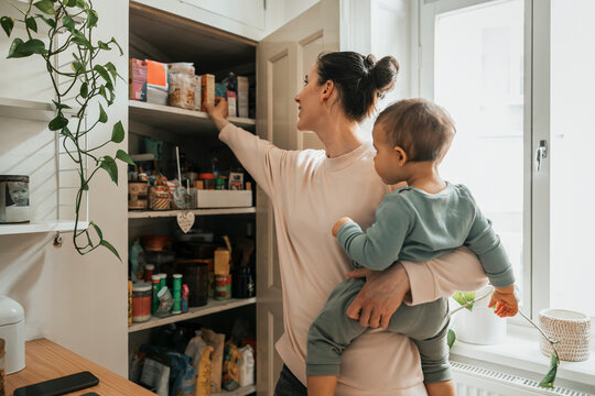 Mother carrying baby boy removing ingredient from pantry in kitchen at home