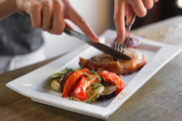 Girl cuts a cooked pork steak served on a white plate with grilled vegetables. Young woman using knife and fork to cutting meat eating in a restaurant. Balanced diet, cooking, culinary. Food concept