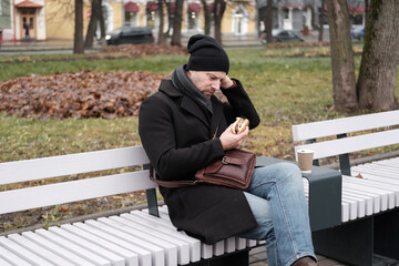 Pensive hipster drinking coffee sitting on a bench in the urban park. Handsome man wearing black...