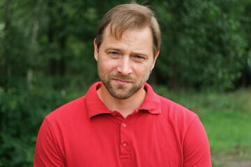 Portrait of handsome young man in a red t-shirt outdoors. Male with fair hair and beard feel happy,...