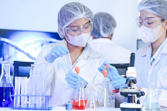 A diverse team of scientists collaborates in a genetics lab. A male researcher explains a procedure with a liquid sample while a DNA helix is displayed on a monitor behind them.