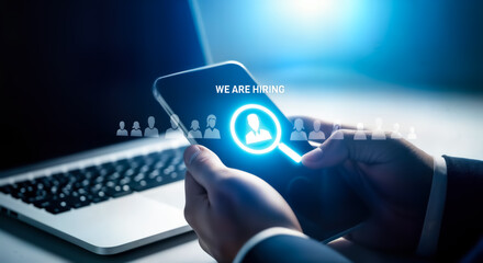 Closeup of a person holding a smartphone with a we are hiring message and a magnifying glass icon, symbolizing recruitment and job search, with a laptop in the background