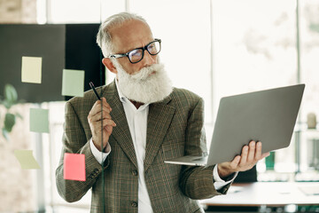 Confident senior businessman working in office with laptop and sticky notes, demonstrating...