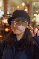 Young woman wearing black cap and navy hoodie playfully looks through magnifying glass in vintage store with warm ambient lighting
