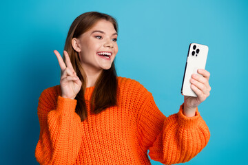 Charming young woman taking a selfie with peace gesture wearing an orange sweater against a blue...