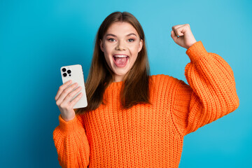 Cheerful young woman holding smartphone in casual stylish outfit celebrating success on blue solid...