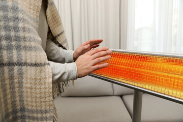 Man with plaid warming his hands near electric heater at home, closeup