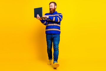 Fototapeta premium Stylish man with a beard holding a laptop while walking against a vibrant yellow background in casual fashion