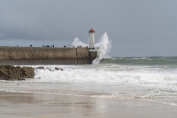 Un phare sur une jet&eacute;e battue par de puissantes vagues sous un ciel orageux, avec l&rsquo;oc&eacute;an en mouvement et une plage au premier plan.