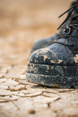 A heavily worn black boot covered in dried mud rests on parched, cracked earth. The desert ground shows unique fissures, suggesting a rugged environment at Burning Man festival.