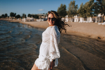 Photo, portrait of curly brunette, beautiful happy smiling young woman walking, walking in sunglasses on the water in the sea, ocean on the beach at sunset in a white sundress, dress.