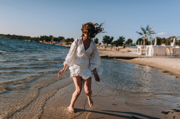 Photo, portrait of curly brunette, beautiful happy smiling young woman walking, walking in sunglasses on the water in the sea, ocean on the beach at sunset in a white sundress, dress.