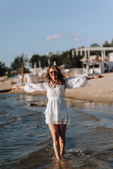 Photo, portrait of a beautiful happy young woman walking, walking in sunglasses on the water in the sea, ocean on the beach at sunset in a white sundress.
