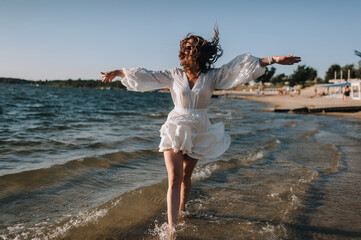 Photo, portrait of a beautiful happy young woman walking, walking in sunglasses on the water in the sea, ocean on the beach at sunset in a white sundress.